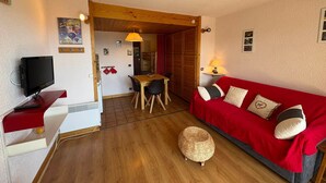 A compact living space with a wooden floor, a red sofa, a small table, a television, and a kitchenette area. - 1-star studio-cabin at Les Saisies, 4 people, south balcony (Les Saisies)