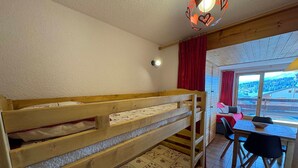 A bunk bed with a patterned quilt, a dining table with chairs, and a view of a landscape through a window. - 1-star studio-cabin at Les Saisies, 4 people, south balcony (Les Saisies)