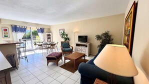 A living room with a sofa, armchair, and coffee table, a TV, and a balcony with a dining area.