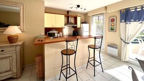 A compact kitchen with a bar area, a microwave, a stove, and a refrigerator. There are two bar stools and a small table with a lamp.