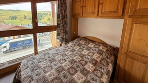 A bedroom with a wooden ceiling, a bed with a patterned quilt, wooden cabinets, and a window with a view of a road and buildings. - Les Saisies - Spacious 3 Rooms Mezzanine 2 Bathrooms for 8 Pers. (Les Saisies)