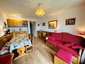 A cozy living space with a dining area, a red patterned sofa, wooden furniture, and a kitchen with wooden cabinets.