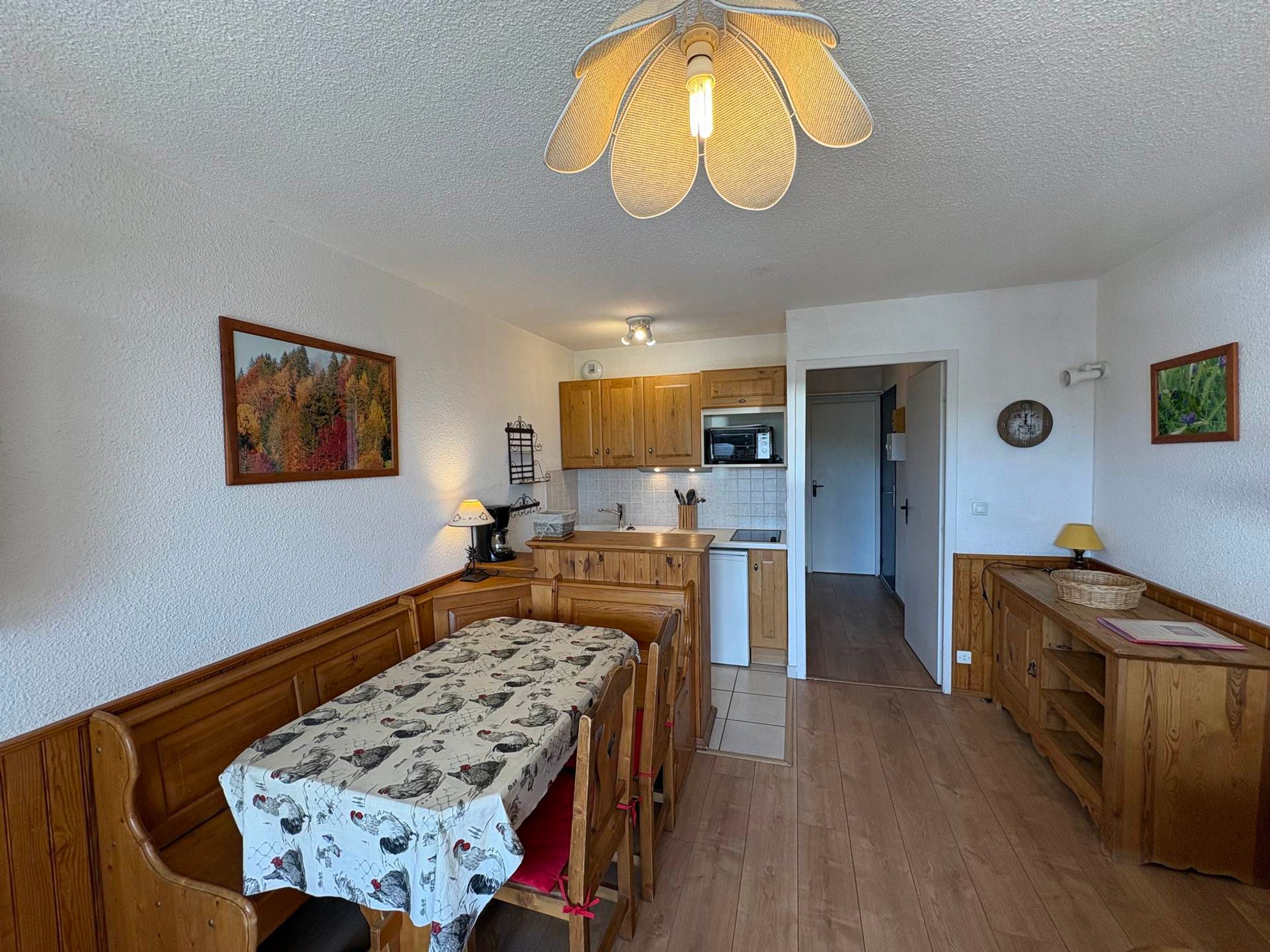 A compact kitchen with wooden cabinets, a dining table with a patterned tablecloth, and a hanging light fixture.