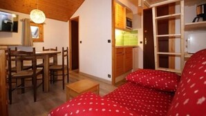 Une pièce chaleureuse avec un plafond en bois, une table à manger avec des chaises, un lit avec une literie rouge et un coin cuisine doté de meubles en bois et d’un four à micro-ondes.