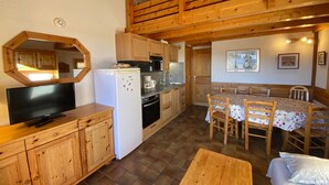 Une cuisine compacte avec des meubles en bois, un réfrigérateur blanc, un four à micro-ondes et une machine à café. On y trouve une table à manger avec des chaises et un téléviseur posé sur un meuble en bois.