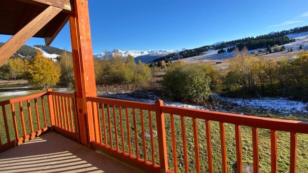 Un balcon en bois avec une balustrade rouge, donnant sur un paysage montagneux aux sommets enneigés et aux arbres aux couleurs automnales.