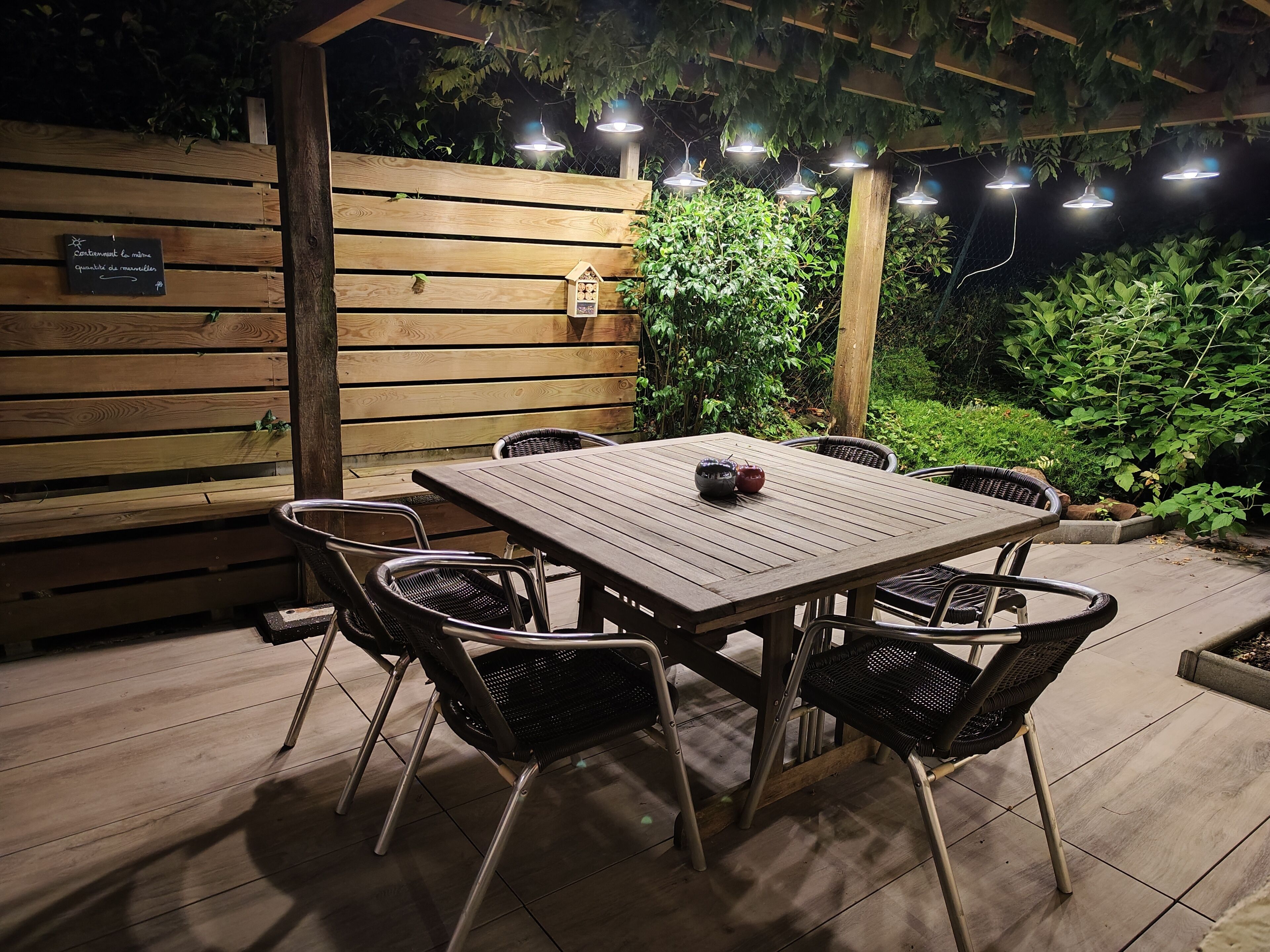 An outdoor patio with a wooden table and chairs, surrounded by greenery and string lights.