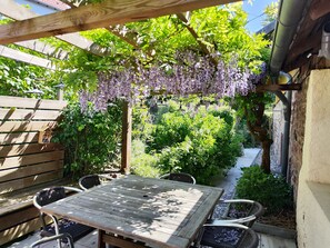 Puinen pergola, jossa on violetteja kukkia, sekä puutarha-alueella oleva pöytä ja tuolit.