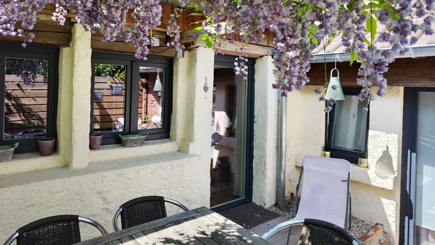 An outdoor patio with a table and chairs, a hanging light fixture, and purple flowers.