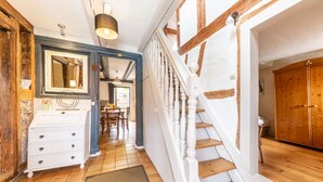 A hallway with a staircase, wooden beams, a white dresser, and a dining area in the background.