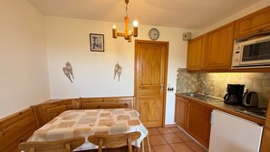 A compact kitchen with wooden cabinets, a dining table with chairs, a clock, and a microwave. - Cozy studio in Les Saisies, 5 pers, south balcony (Les Saisies)