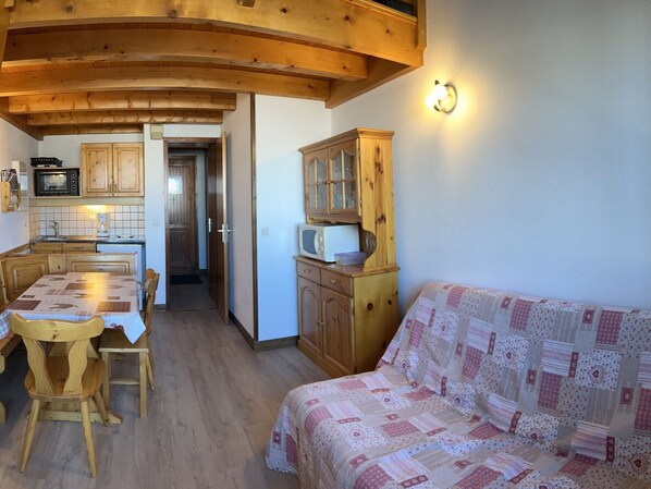 A cozy room with a wooden ceiling, a dining table with chairs, a quilted sofa, and a kitchen area with wooden cabinets and a microwave.