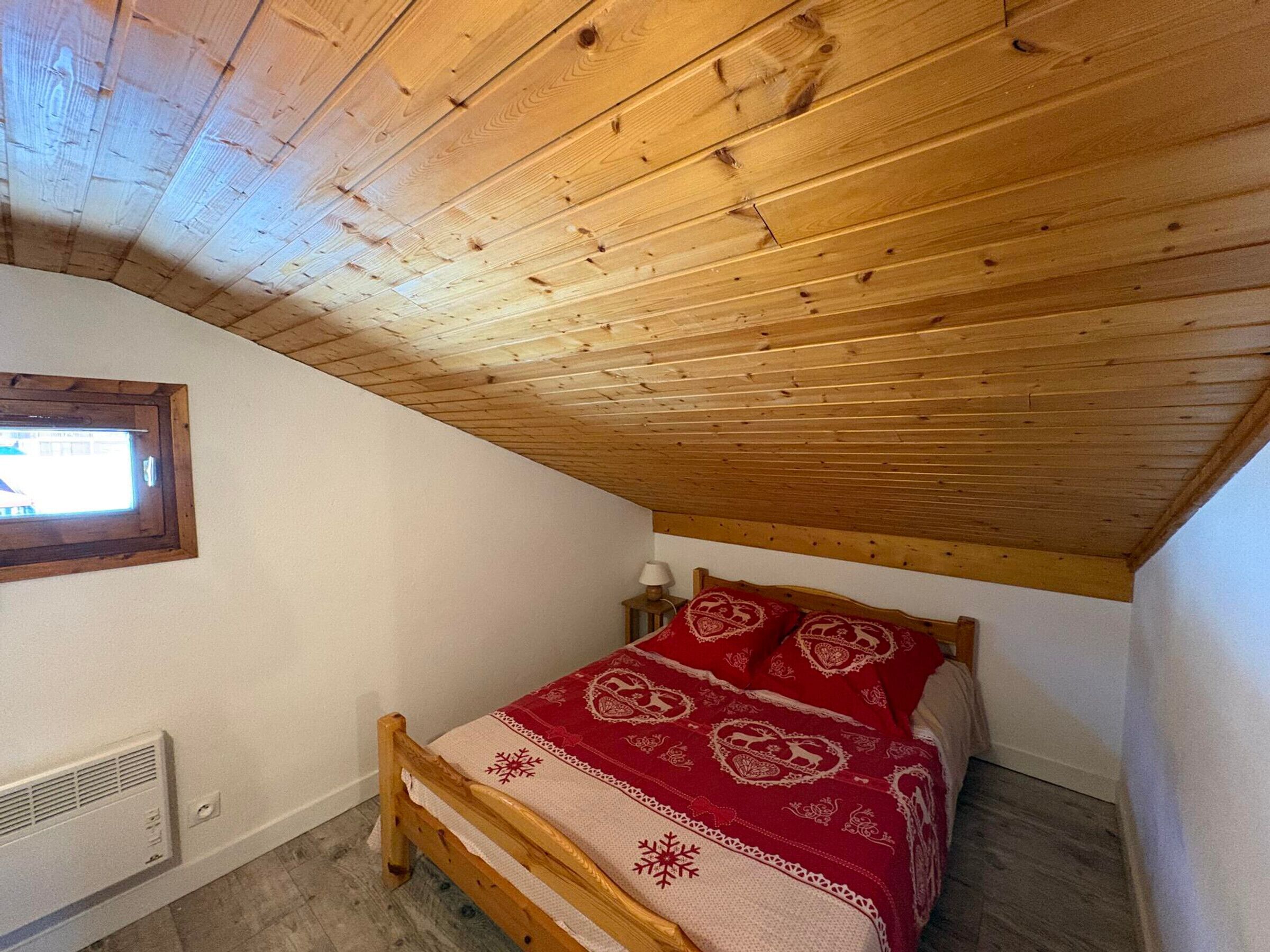 A bedroom with a wooden ceiling, a bed with a red patterned bedspread, a window, and a wall-mounted heater.