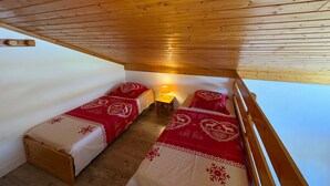 Une petite pièce chaleureuse avec un plafond et des murs en bois, deux lits simples avec une literie rouge et blanche, et une petite table avec une lampe.