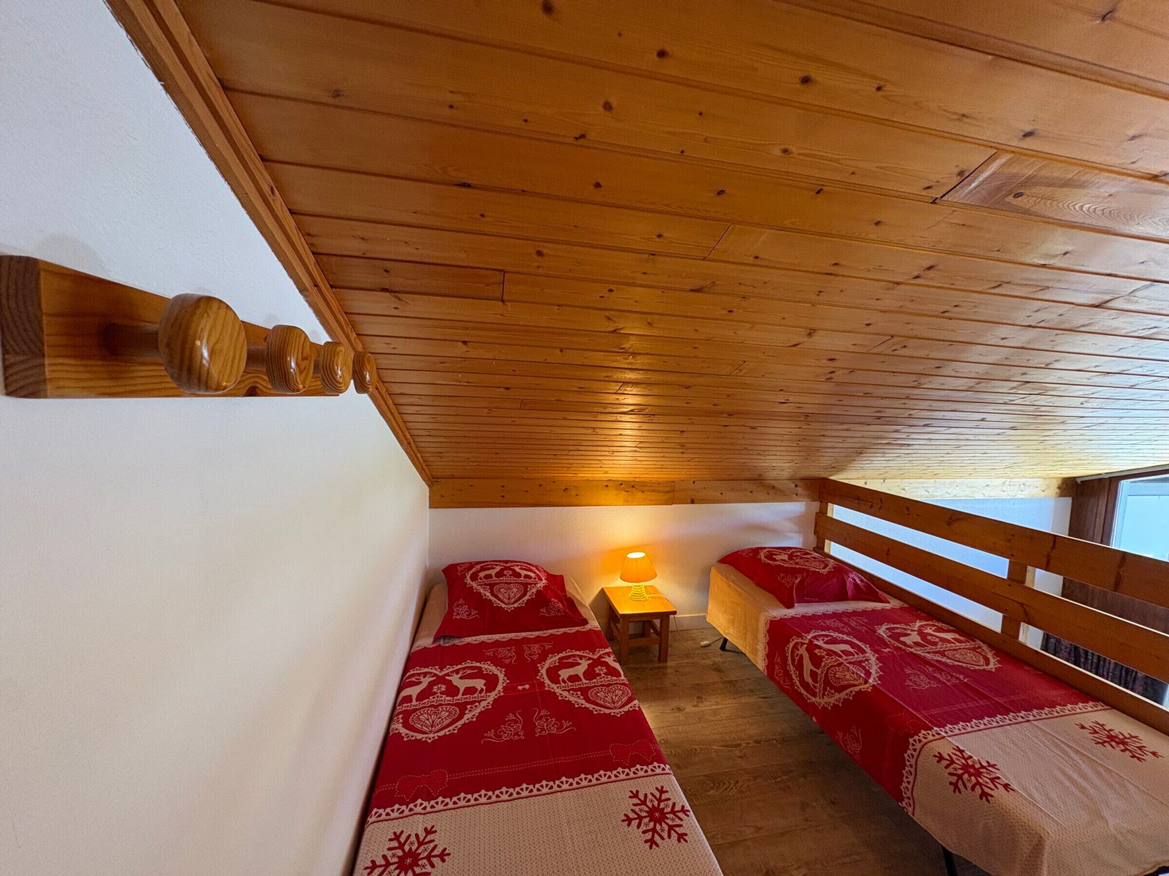 A small, cozy room with wooden ceiling beams, two beds with red and white bedding, a small table with a lamp, and a wooden railing.