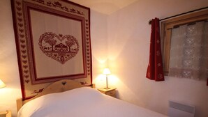 A neatly made bed with a heart-shaped tapestry on the wall, a bedside lamp, and a window with a curtain. - Cozy apartment in Les Saisies - 4/6 pers, 38m², south, balcony (Hauteluce)