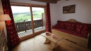 A cozy living room with a wooden floor, a sofa with red cushions, a small table, and a window with a view of a green landscape. - Cozy apartment in Les Saisies - 4/6 pers, 38m², south, balcony (Hauteluce)