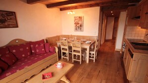 A cozy living room with a wooden sofa, a dining table, and chairs. A kitchen with wooden cabinets and a sink is visible in the background. - Cozy apartment in Les Saisies - 4/6 pers, 38m², south, balcony (Hauteluce)