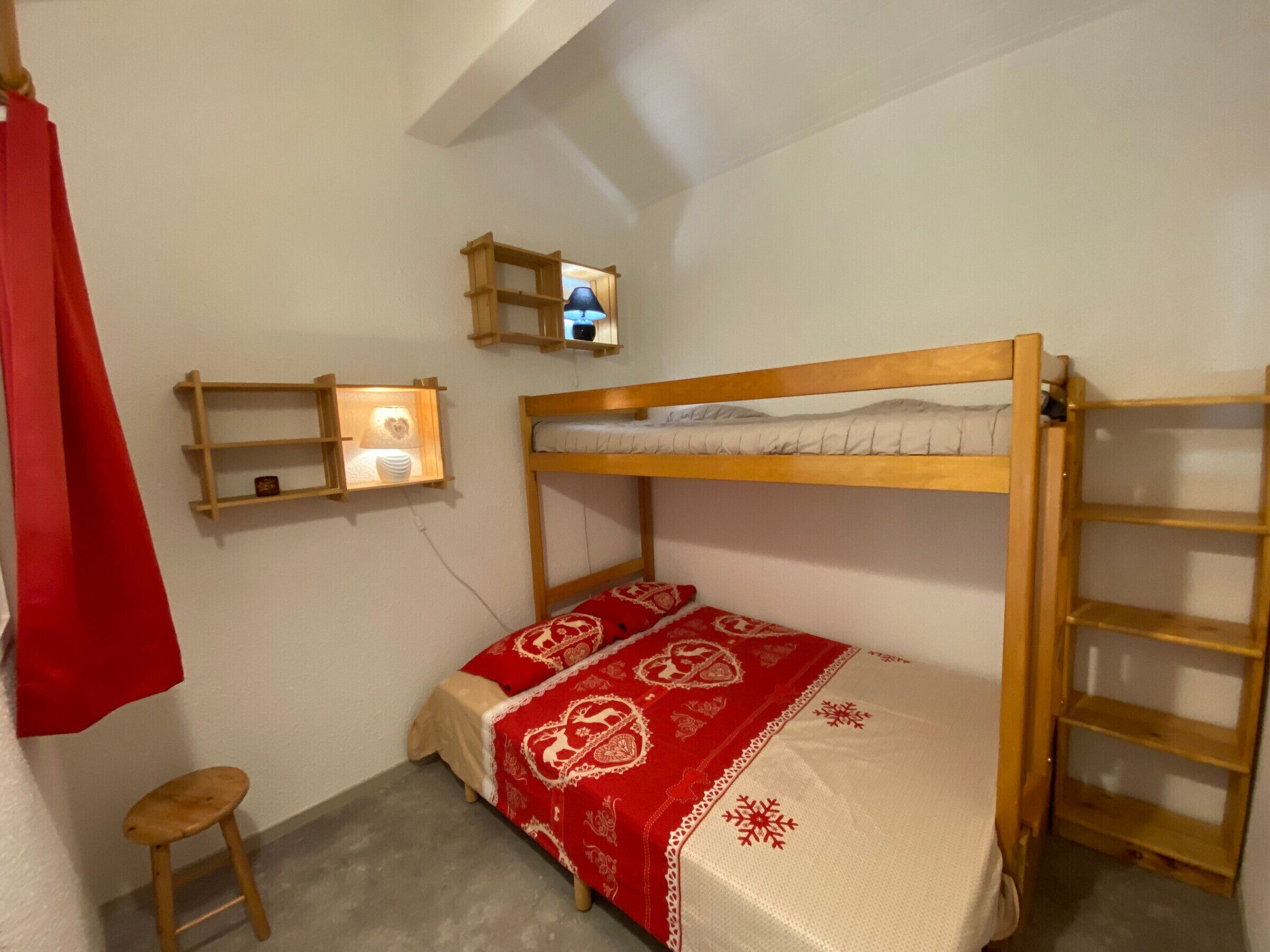 Un letto a castello con copripiumino rosso e bianco a motivi, un piccolo sgabello di legno e uno scaffale con una lampada.