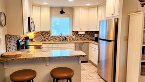 Una cocina con armarios blancos, encimera de granito, refrigerador de acero inoxidable y ventana con persianas.