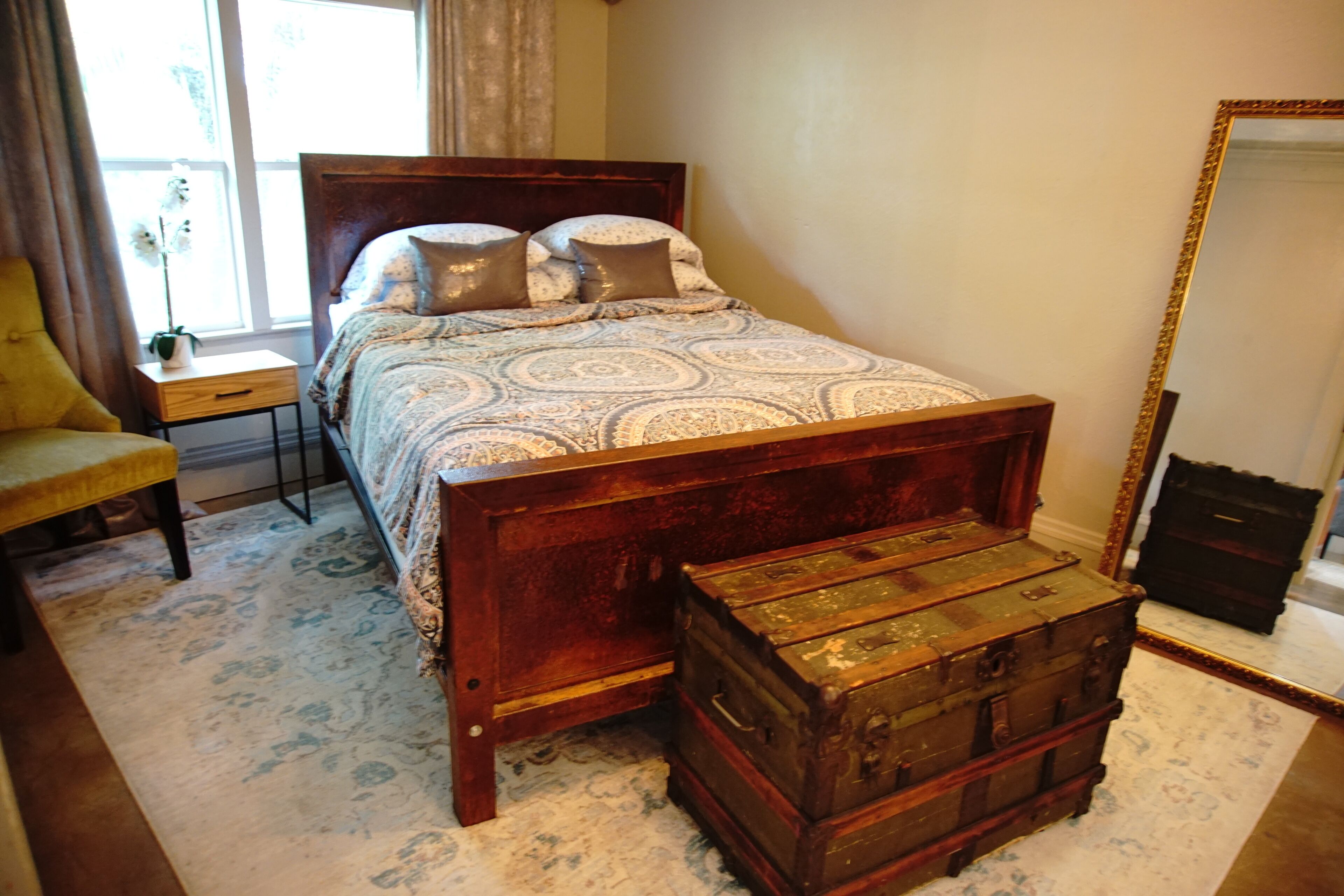 A bedroom with a wooden bed, a nightstand, a chair, a large mirror, and a vintage trunk.