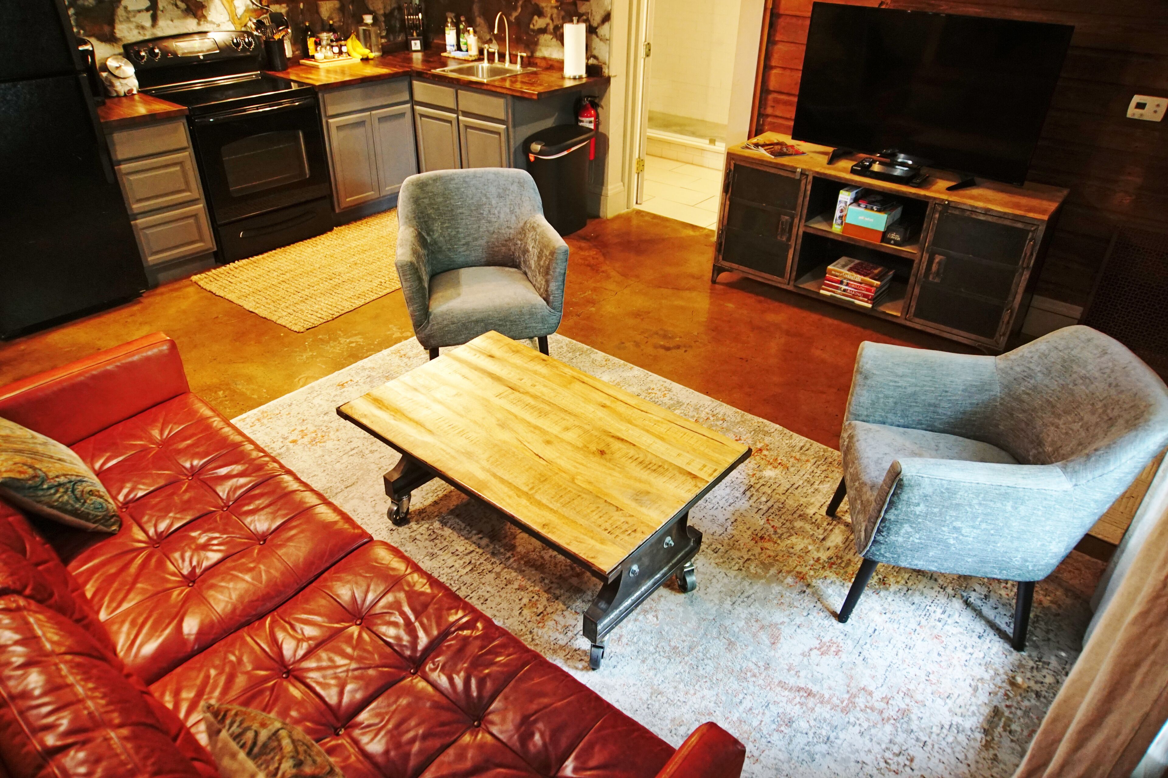 Un salon avec un canapé en cuir rouge, une table basse en bois et deux fauteuils gris. On aperçoit au fond une cuisine avec des meubles sombres et un fourneau.