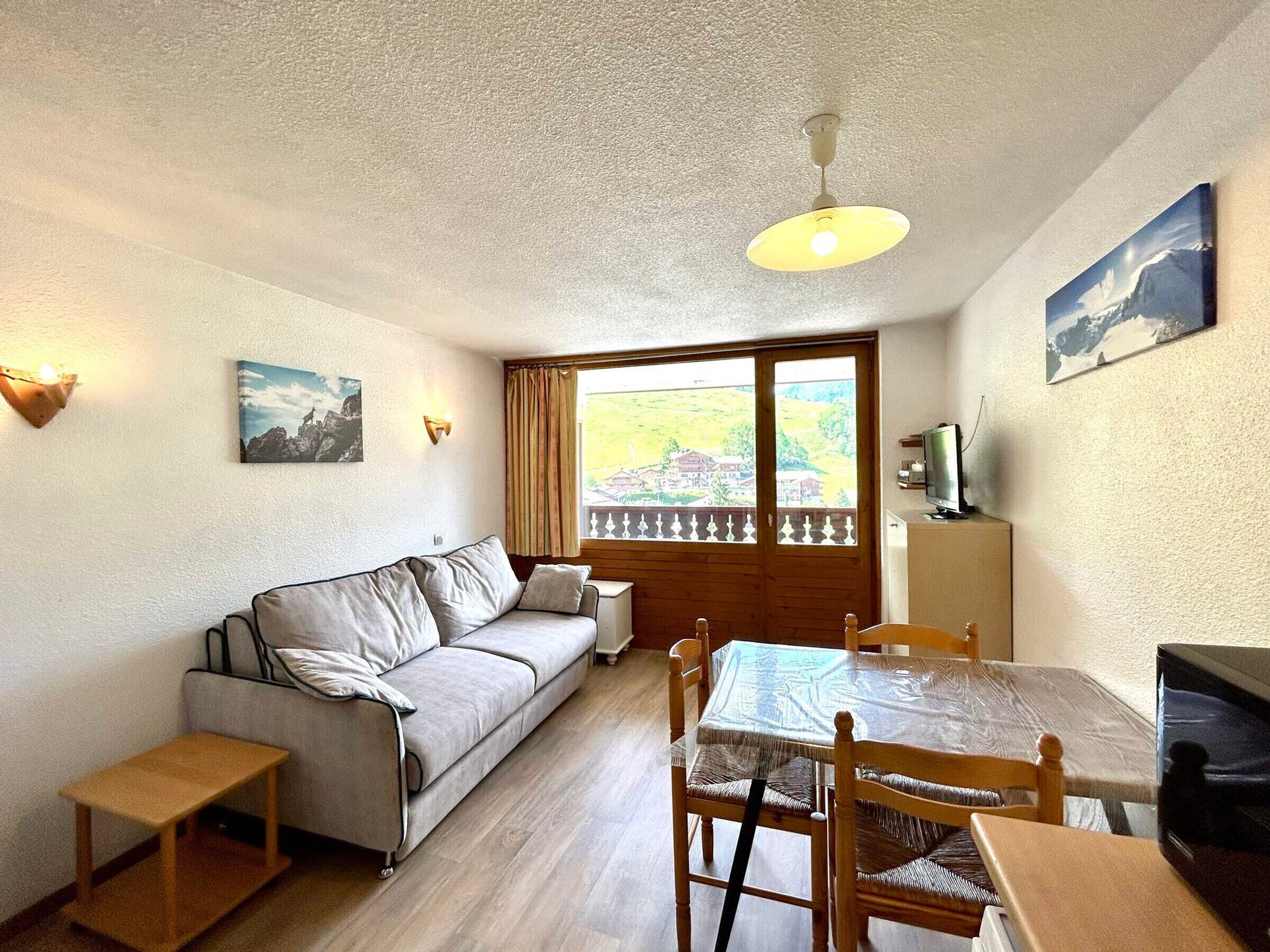 A cozy room with a sofa, a dining table, and chairs. There's a window with a view of a house and greenery.