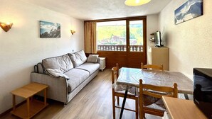 A cozy room with a sofa, a dining table, and chairs. There's a window with a view of a house and greenery. - Studio for 4 people with garage, near village and lifts (La Clusaz)