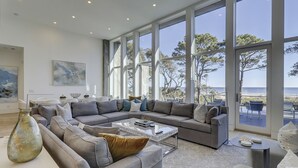En rummelig stue med en stor grå hjørnesofa, et sofabord og havudsigt gennem store vinduer.