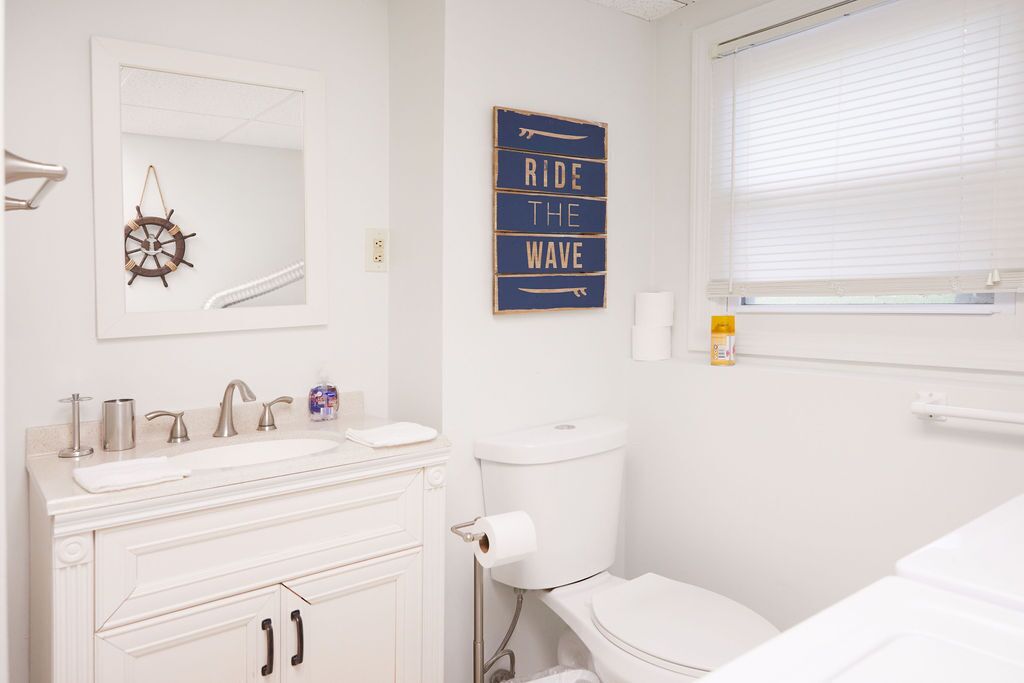 A bathroom with a white sink, vanity, and toilet. There is a mirror above the sink and a sign on the wall that says "RIDE THE WAVE".