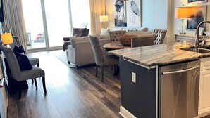 Een moderne keuken met een centraal eiland, een eethoek met een bank, en een woonkamer met een groot raam en gordijnen.