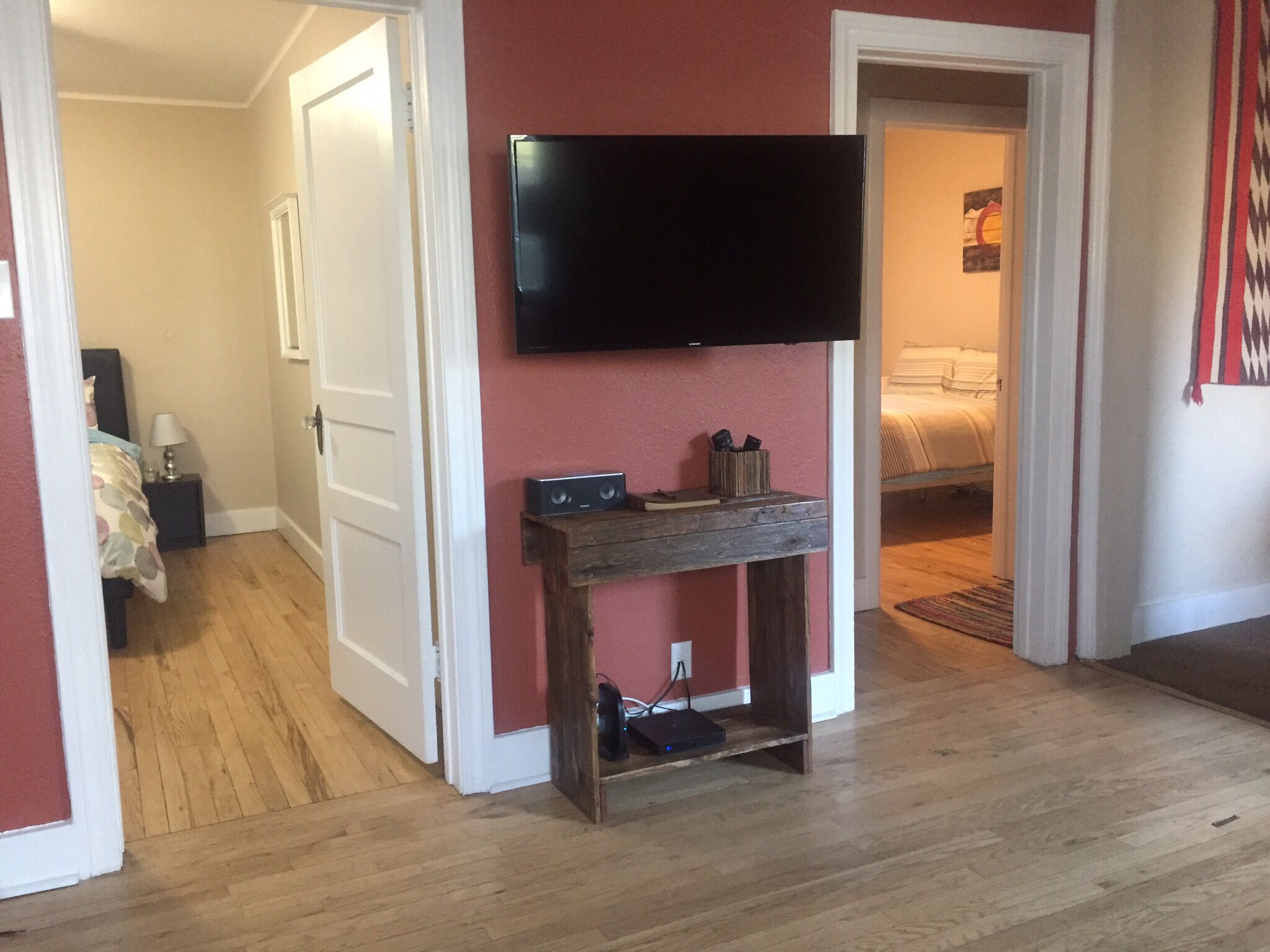 Una habitación con piso de madera, un televisor de pantalla plana montado en la pared, una pequeña mesa consola de madera con un altavoz y libros, y una cama con cubrecama blanco al fondo.