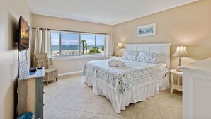 Un dormitorio con una cama grande, un televisor montado en la pared, una ventana con vistas a la playa y una mesita de noche con una lámpara.