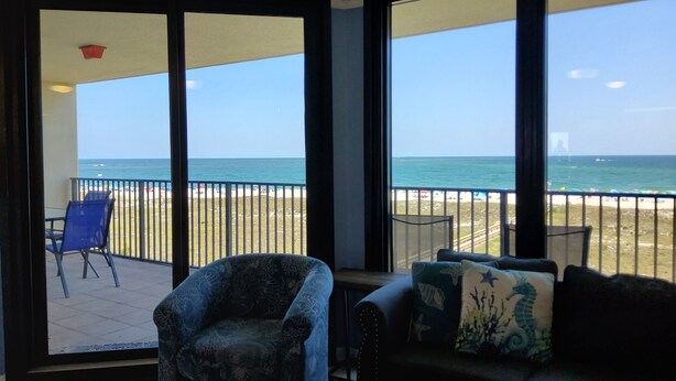Ein Zimmer mit Blick auf den Strand und den Ozean durch große Fenster. Ein blauer Sessel und eine Couch mit dekorativen Kissen sind vorhanden.