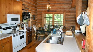 A log cabin kitchen with wooden cabinets, a white stove, a sink, and a dining area with chairs and a table.