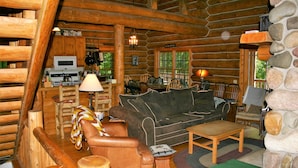 A log cabin interior with a wooden dining area, a sofa, a leather armchair, and a stone wall.