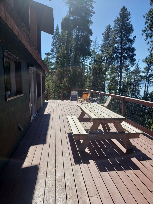 A wooden deck with a picnic table and chairs, surrounded by tall trees. - Peaceful home enveloped in a quiet, scenic landscape adjacent to a rocky beach. (Bigfork)