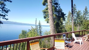 Une terrasse en bois avec quatre chaises et une table, donnant sur un lac bordé d’arbres et sur des montagnes lointaines.