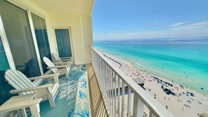 Un balcon avec des chaises blanches et un sol à motifs, donnant sur une plage où l’on aperçoit des gens et des parasols.