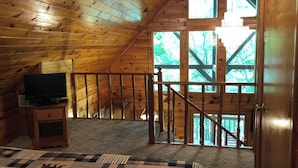 A wooden cabin interior with a TV on a stand, a staircase leading to a balcony, and a view of trees through the windows. - *Winter Special* 2 bed 2 bath 10 min PF Private (Sevier County)