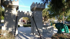 A stone archway with chains, leading to a pathway surrounded by greenery and palm trees.