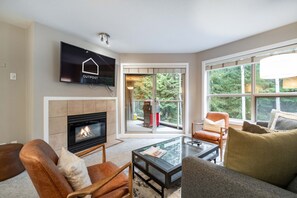 A modern living room with a fireplace, comfortable seating, a television, and a large window offering a view of the outdoors. - Ski-in/out | Pool, hot tub, gym | BBQ | Fireplace (Whistler)