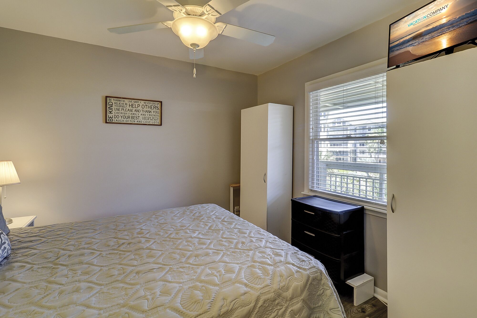 A bedroom with a bed, a ceiling fan, a window with blinds, a wardrobe, and a motivational quote on the wall.