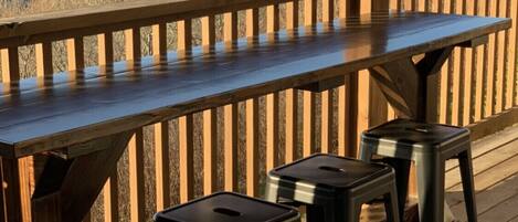 A wooden deck with a long table and three black stools. The deck overlooks a mountainous landscape.