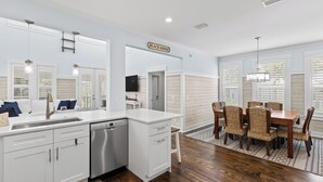 A modern kitchen with white cabinetry, a dishwasher, and a dining area with wicker chairs and a wooden table. - Seacrest's Forever & Always + Pool, Tram & Bikes (Seacrest)