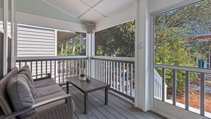 A covered porch with a sofa and table, large windows, and a view of trees and a house. - Seacrest's Forever & Always + Pool, Tram & Bikes (Seacrest)