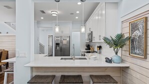 A modern kitchen with a central island, stainless steel appliances, pendant lighting, and a wooden accent wall. - Seacrest's Forever & Always + Pool, Tram & Bikes (Seacrest)