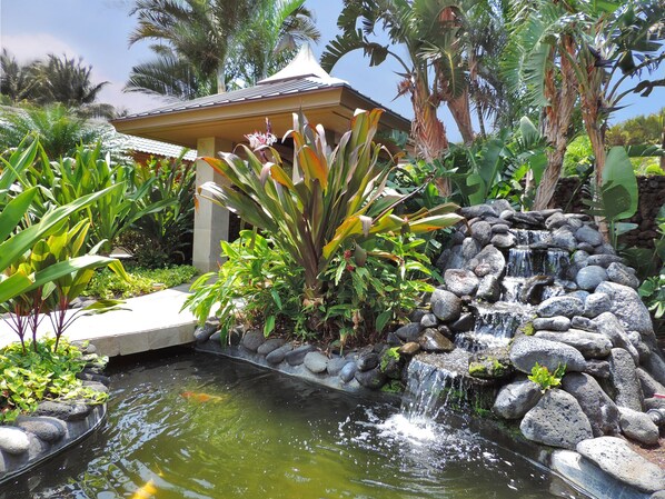 A tranquil garden with a small waterfall, lush tropical plants, and a gazebo.