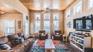 Un salon chaleureux avec un plafond en bois, un ventilateur de plafond, une grande télévision, un tapis coloré, un canapé, deux fauteuils et une bibliothèque remplie de jouets.