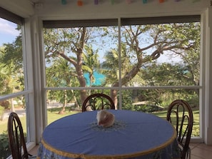 Enjoy a serene view of the lush garden from the large window, which offers a glimpse of a palm tree. The room features a round table with a tablecloth and a single chair.